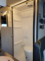 Compact RV rental wet bath interior featuring a toilet, shower, mirror, LED lighting, and essential controls.