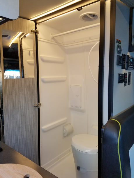 Compact RV rental wet bath interior featuring a toilet, shower, mirror, LED lighting, and essential controls.