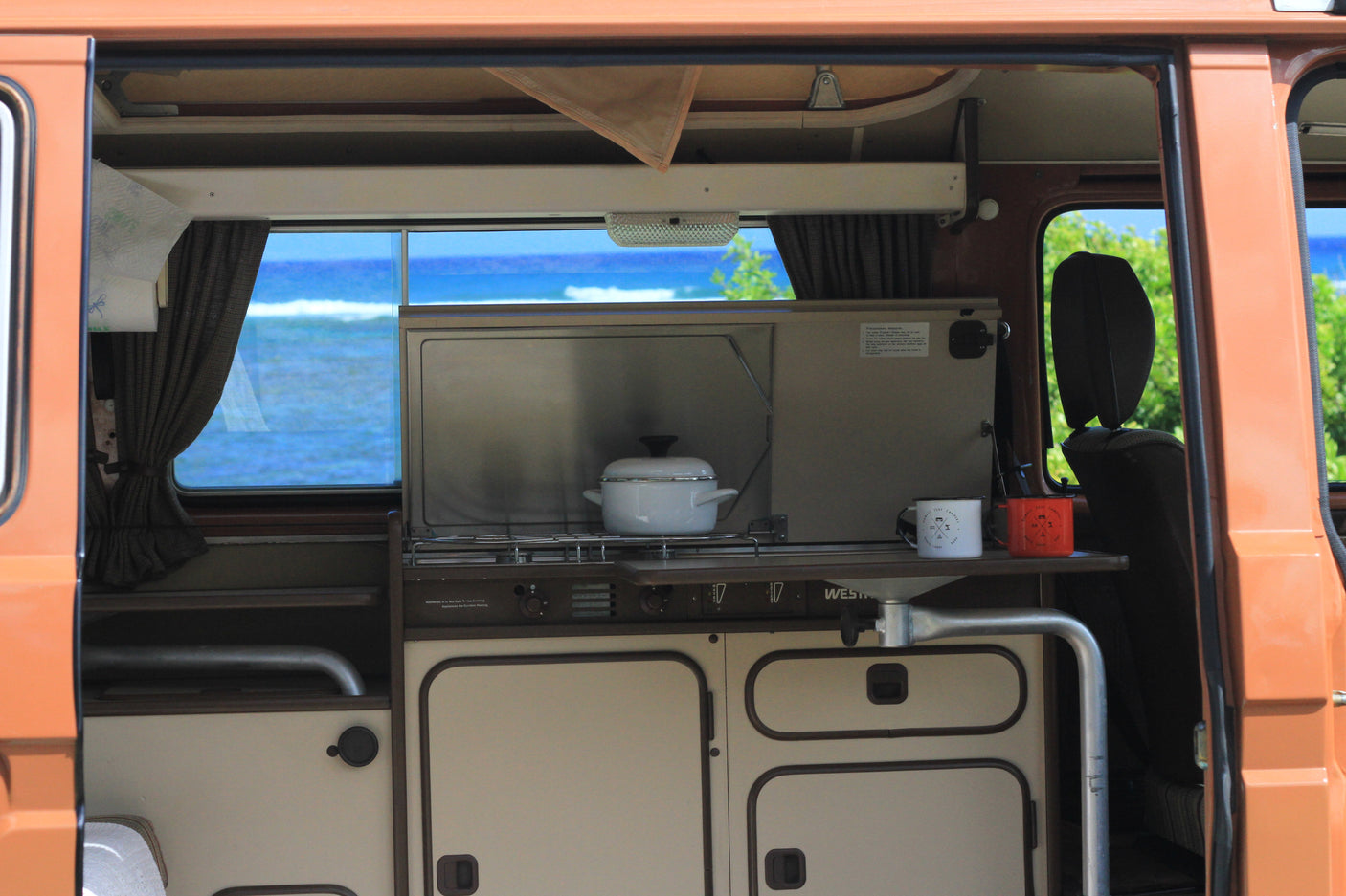 RV rental interior: kitchen with pot & mugs, framed by a stunning ocean view & curtains.