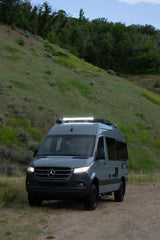 A gray Mercedes-Benz Sprinter RV rental van with lights on, parked on a dirt road beside a grassy, wooded hill.