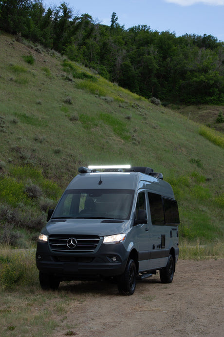 A gray Mercedes-Benz Sprinter RV rental van with lights on, parked on a dirt road beside a grassy, wooded hill.