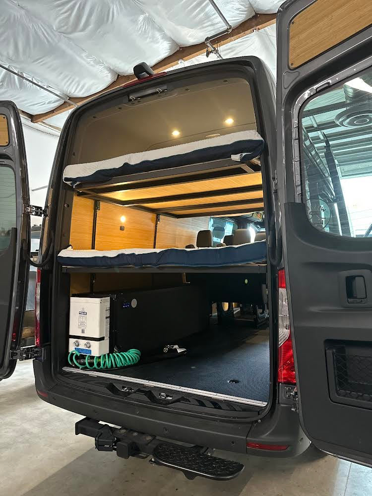 Rear view of an RV rental van's organized interior: bunk beds, a water heater, and storage for gear. Ideal for trips.