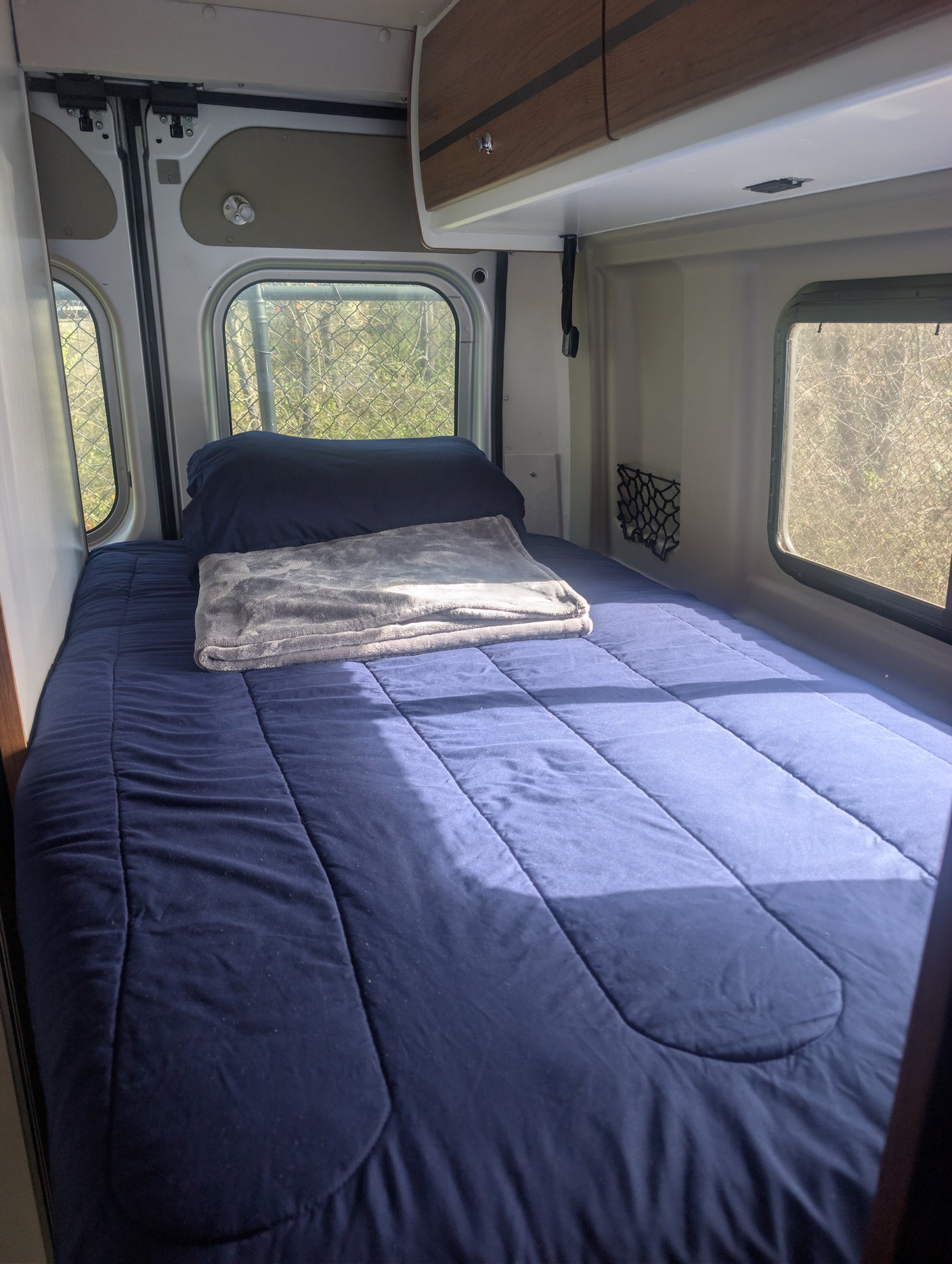 Comfortable bed setup in an RV rental, featuring blue bedding, a grey blanket, and sunlit windows for peaceful travels.