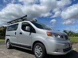 Compact silver Nissan NV200 RV rental camper van with rooftop awning and watersports gear under a sunny sky.