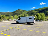 White RV rental van parked alone in a scenic mountain lot. Blue sky & green trees enhance the travel vibe.