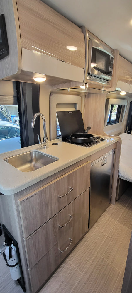 Interior of an rv rental showing a bright, compact kitchen with a sink, stovetop, microwave, fridge, and storage.