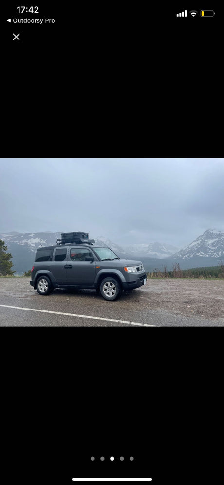 Gray Honda Element with roof box parked on wet pavement, snowy mountains visible under cloudy skies. Plan your next RV rental adventure!