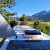 Looking out from an RV rental, showing solar panels on the roof, with a serene view of mountains, a lake, and pine trees.