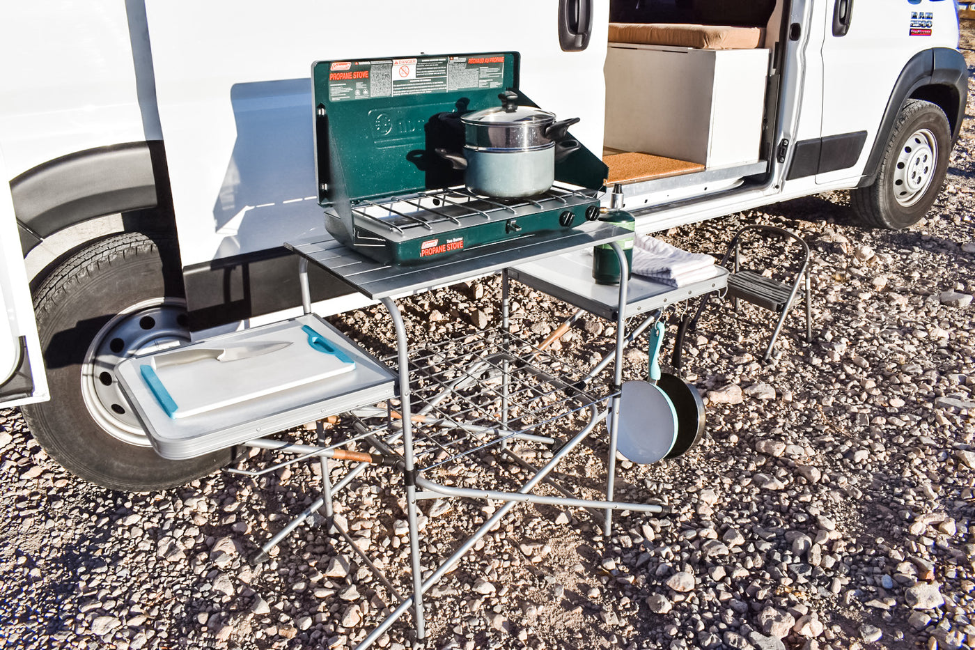 RV rental outdoor kitchen: Coleman stove, cutting board, and pans set up on a portable table beside a camper van. — photo 6