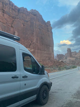 A light-colored camper van with a roof-mounted awning parked on a road next to massive red rock cliffs. Ideal RV rental.