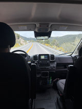 View from an RV rental's passenger seat. Driver steers on a mountain road with navigation active, cloudy skies above.