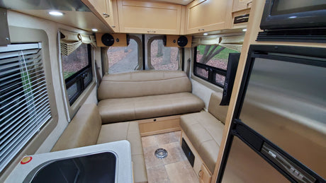 Cozy RV rental interior: tan upholstered seating, wood cabinets, compact kitchen with sink, fridge/microwave. Scenic views from windows.