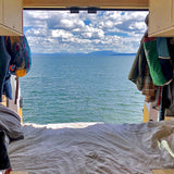 A cozy bed inside a camper van overlooks a calm lake, distant snowy mountains, and cloudy sky. Perfect RV rental escape!