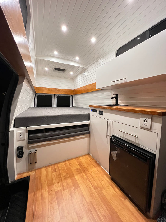 Bright, modern camper van interior for your next RV rental adventure. Features bed, kitchen, fridge, and warm wood accents.