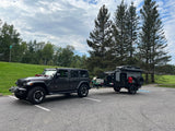 Dark gray Jeep tows a rugged overlanding rv rental trailer in a paved lot, surrounded by green hills & tall trees.