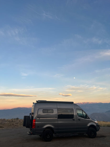 Adventure awaits! A modern gray camper van, ideal for an RV rental, parked with scenic mountain views & moon at dusk.