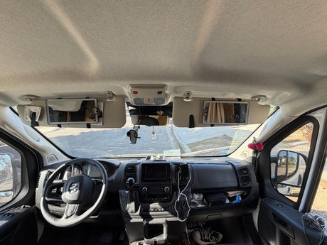 Inside an RV rental: a full view of the front cabin, dashboard, and steering wheel, with sun visors down and personal items visible.