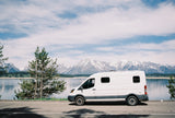 White rv rental campervan parked by a beautiful lake, with majestic snowy mountains in the background. Perfect for outdoor adventures.