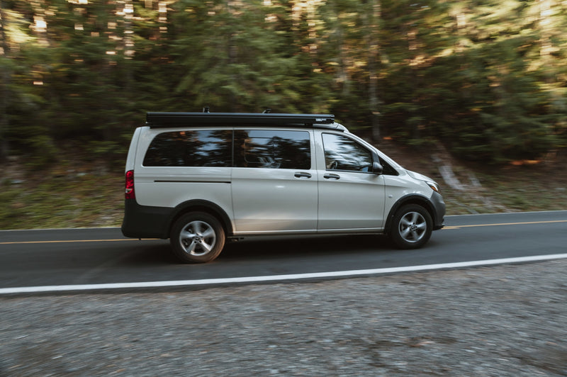  parked by a lake. Surrounded by mountains & evergreen trees under cloudy skies.|RV rental: Light campervan with roof awning on a blurred forest road — photo 2