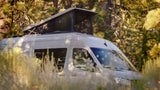 An **RV rental**: A modern campervan with an extended pop-top tent is parked within a dense pine forest for camping.