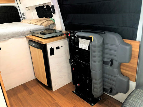 Compact RV rental interior featuring a bed, kitchen with fridge/cooktop, and a foldable seat for versatile living space.