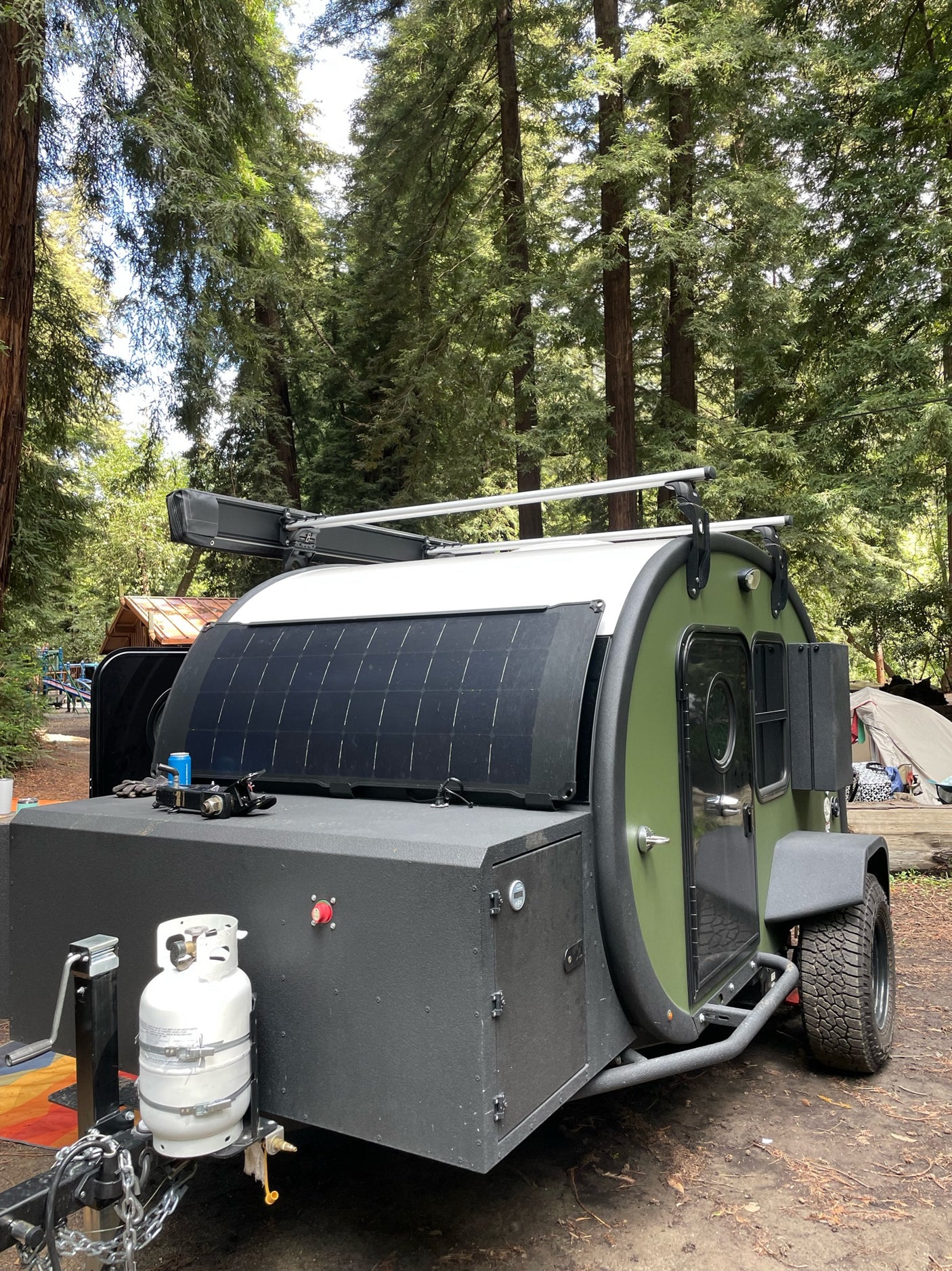 Rugged green off-road teardrop RV rental, with solar panels & propane tank, parked at a tall-treed campground.