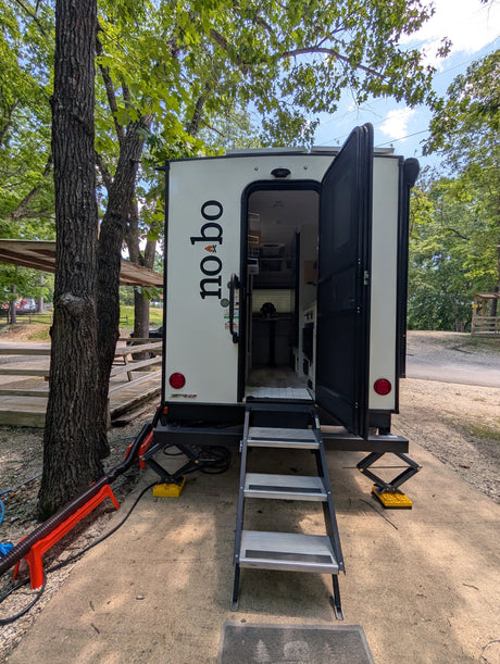 This white "no bo" travel trailer rv rental is set up in a wooded campground, with its door open and steps extended.