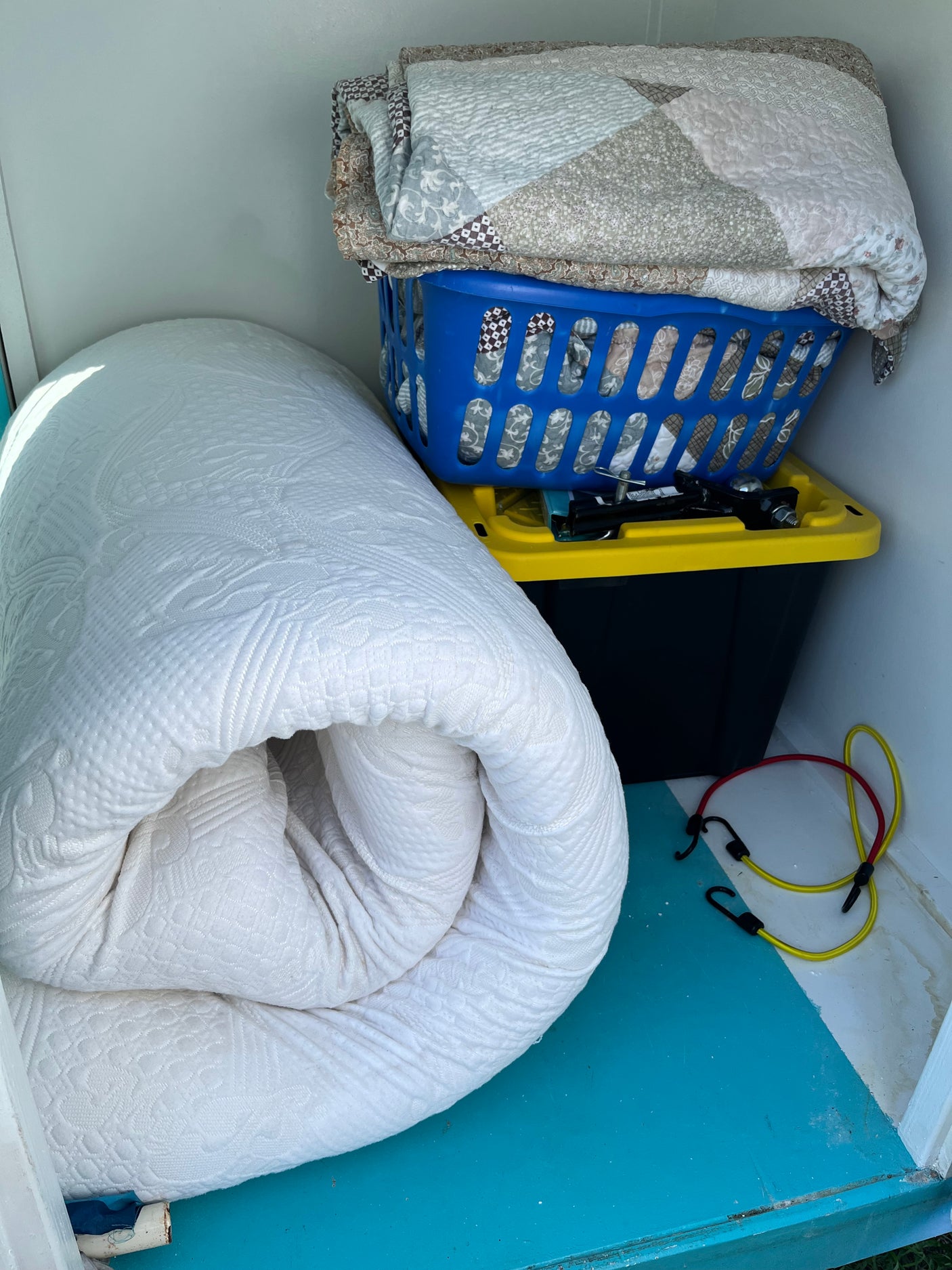 RV rental storage with rolled mattress, blankets in a basket, tools on a bin, and bungee cords on the floor.