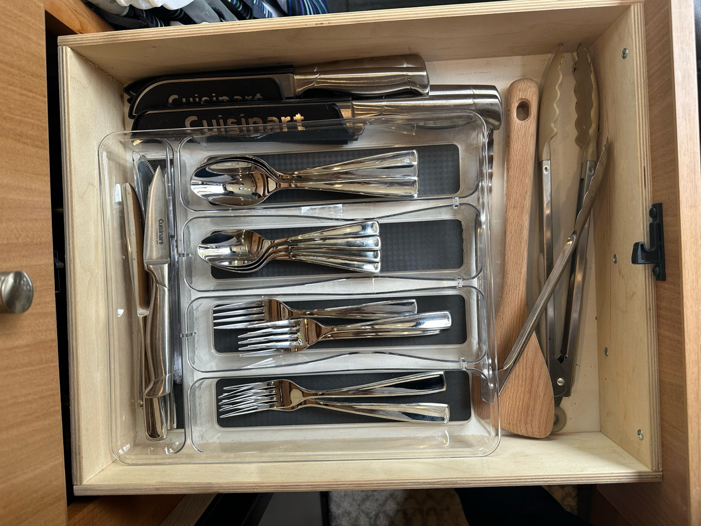 Compact RV rental kitchen drawer: A clear organizer holds shining silverware, a wooden spatula, and tongs.