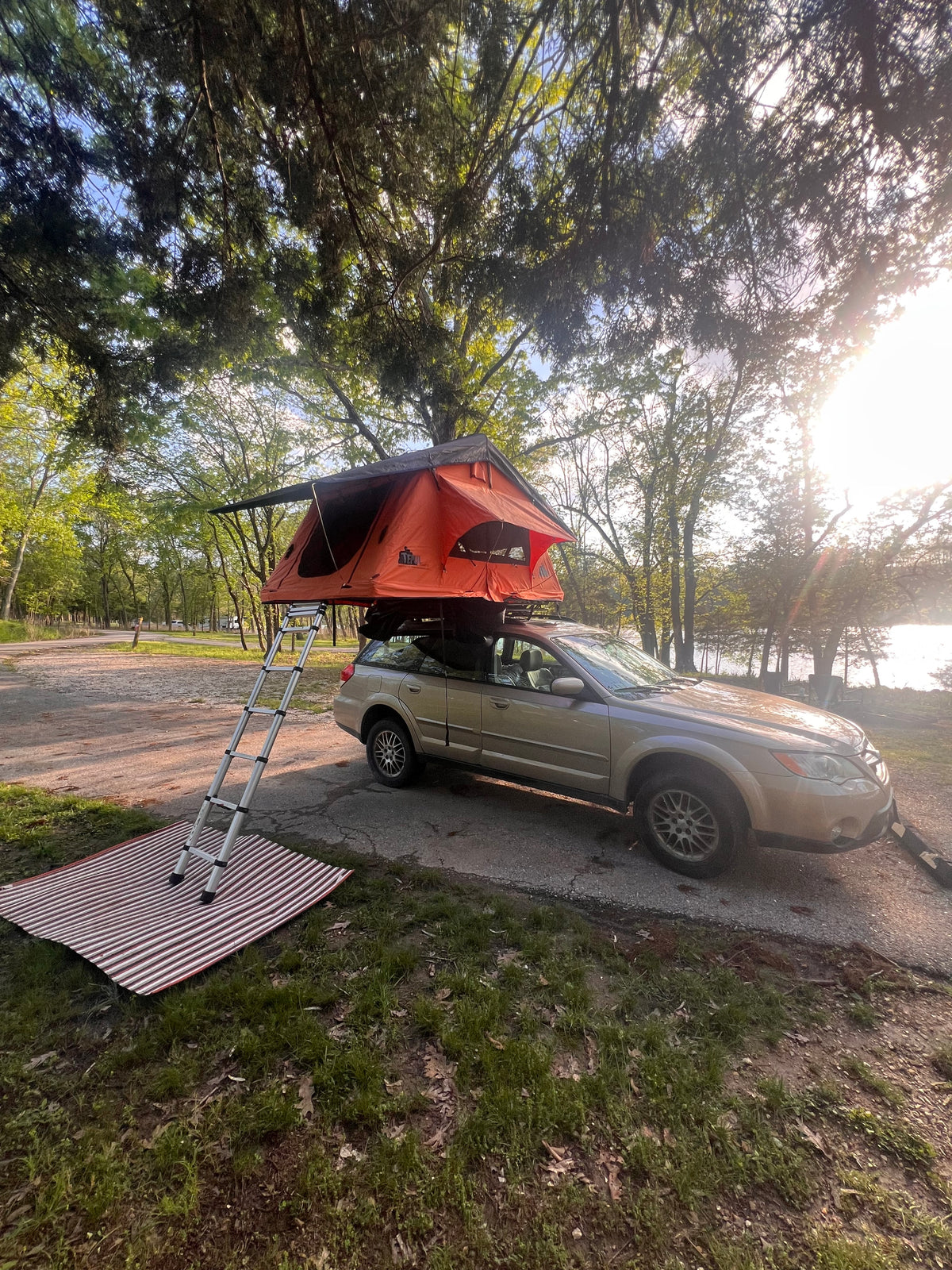 Orange rooftop tent on a Subaru for easy RV rental camping. Setup with ladder and mat at a sunny, wooded lakeside campsite. — photo 3