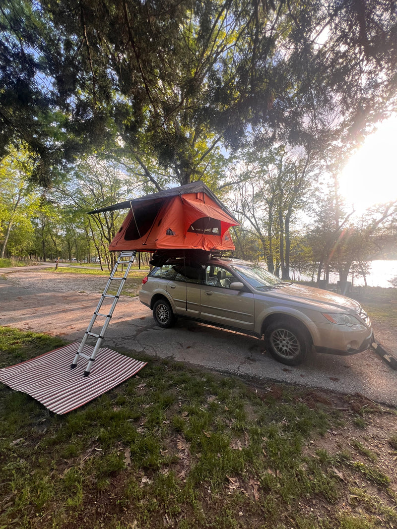 Orange rooftop tent on a Subaru for easy RV rental camping. Setup with ladder and mat at a sunny, wooded lakeside campsite. — photo 3