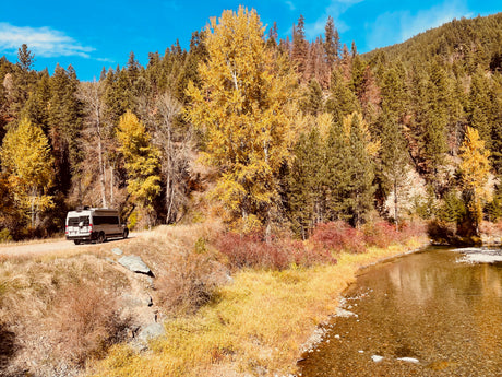 Scenic autumn view with an RV rental on a dirt road beside a clear river and vibrant fall foliage, perfect for nature exploration.