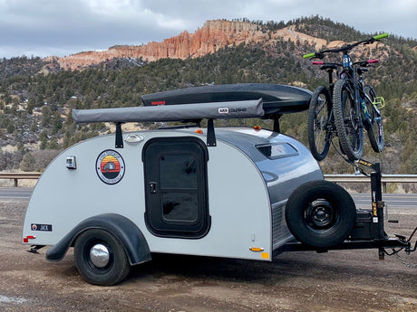An RV rental teardrop camper with bikes and a roof cargo box sits ready for adventure amidst Utah's red rock mountains.