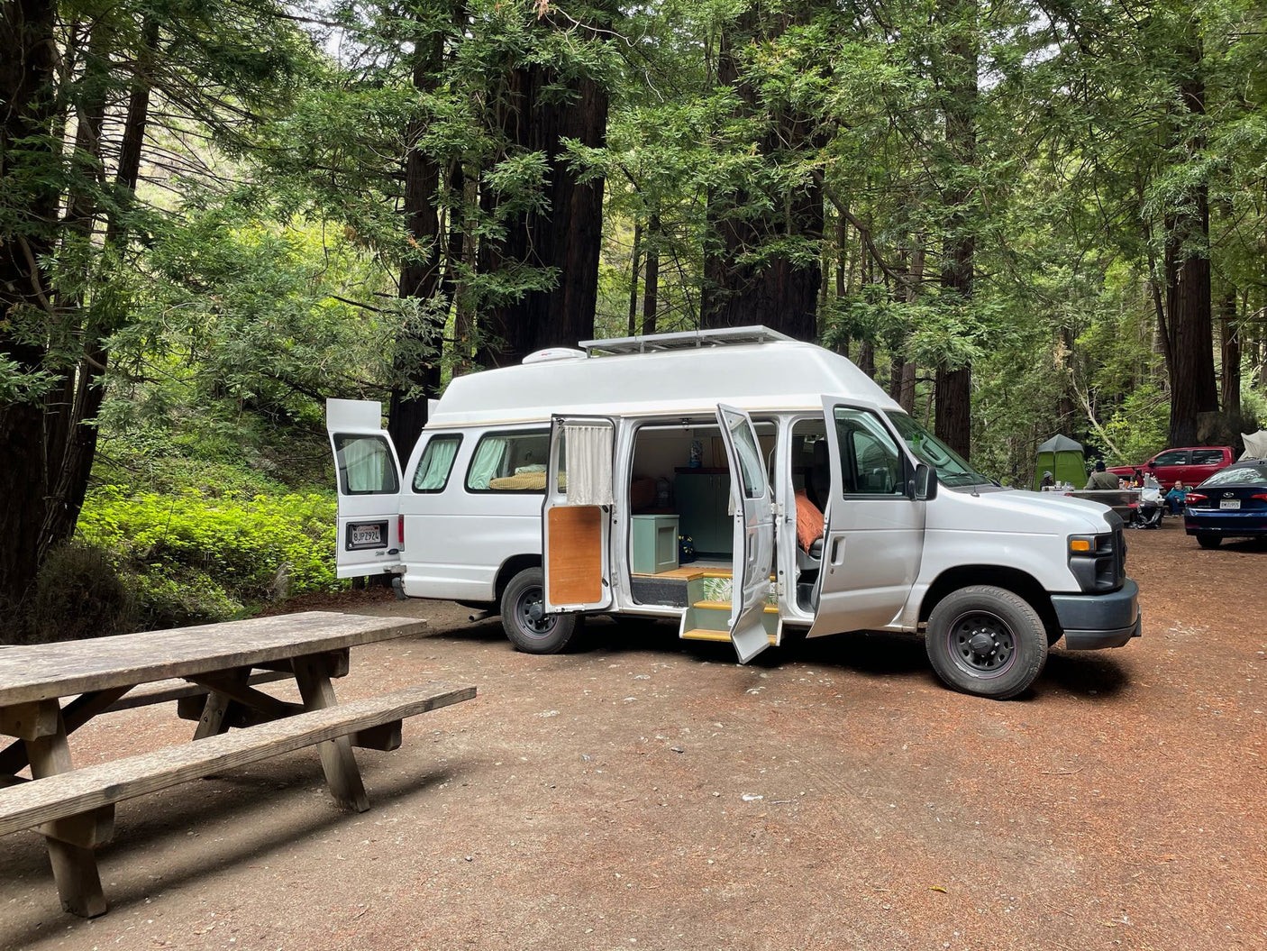 White camper van (RV rental) at a serene redwood forest campsite with open doors, showing interior, beside a picnic table.