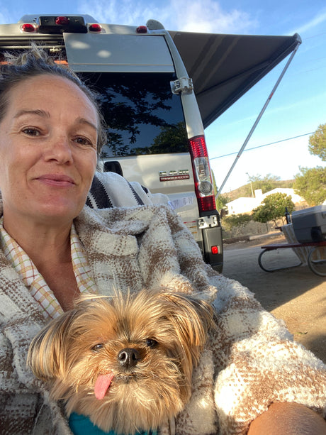 A happy woman and her dog relax in front of an RV rental with its awning extended, enjoying a sunny day outdoors.
