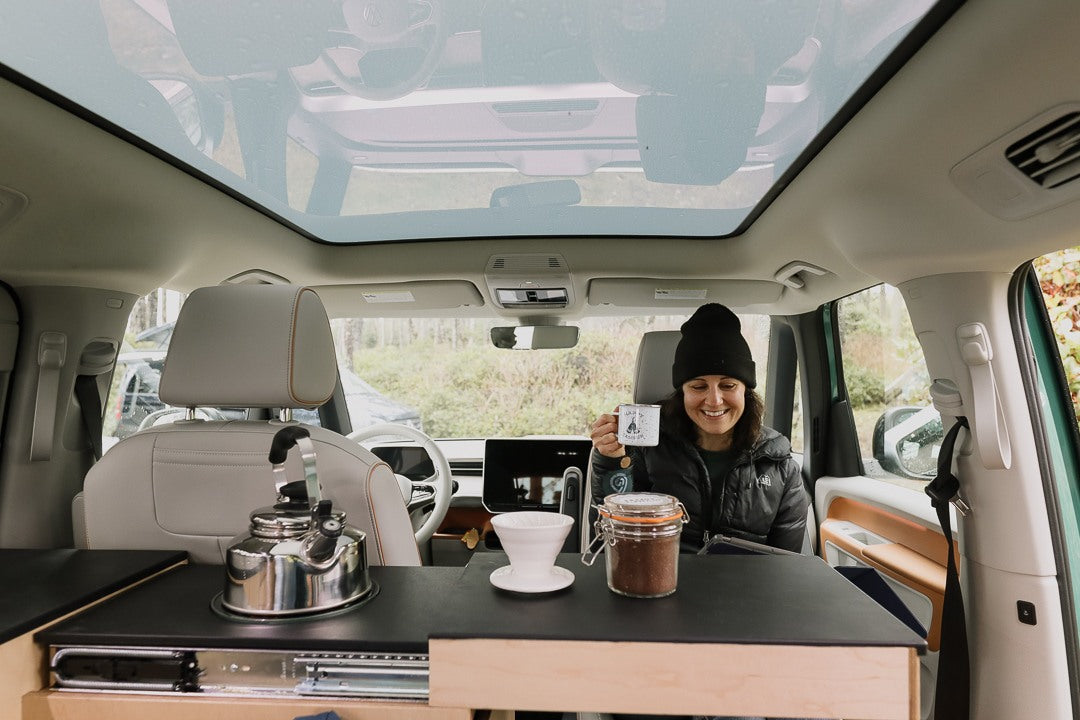 Happy woman making coffee in a modern RV rental camper van, featuring a panoramic sunroof. Perfect for van life adventures.