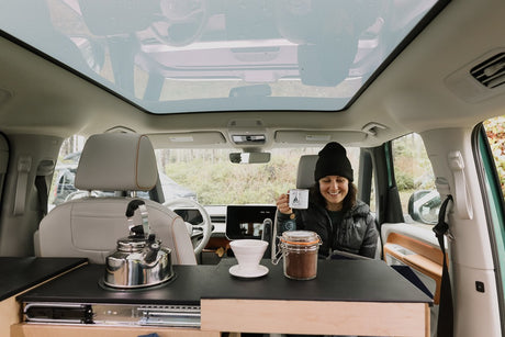 Happy woman making coffee in a modern RV rental camper van, featuring a panoramic sunroof. Perfect for van life adventures.
