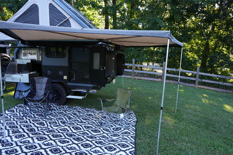 Kick back with this RV rental setup! A modern camper, awning, outdoor kitchen, chairs, and mat on grass for camping.