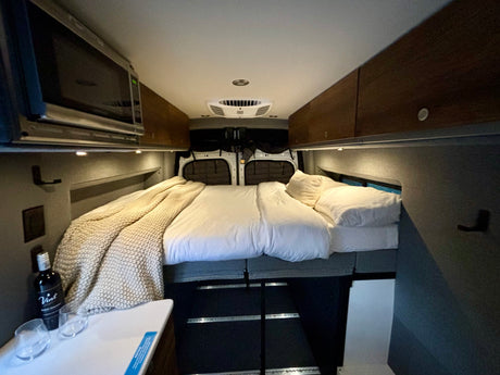 This RV rental features a cozy bed with white linens, overhead storage, a microwave, and wine, offering a comfortable travel interior.