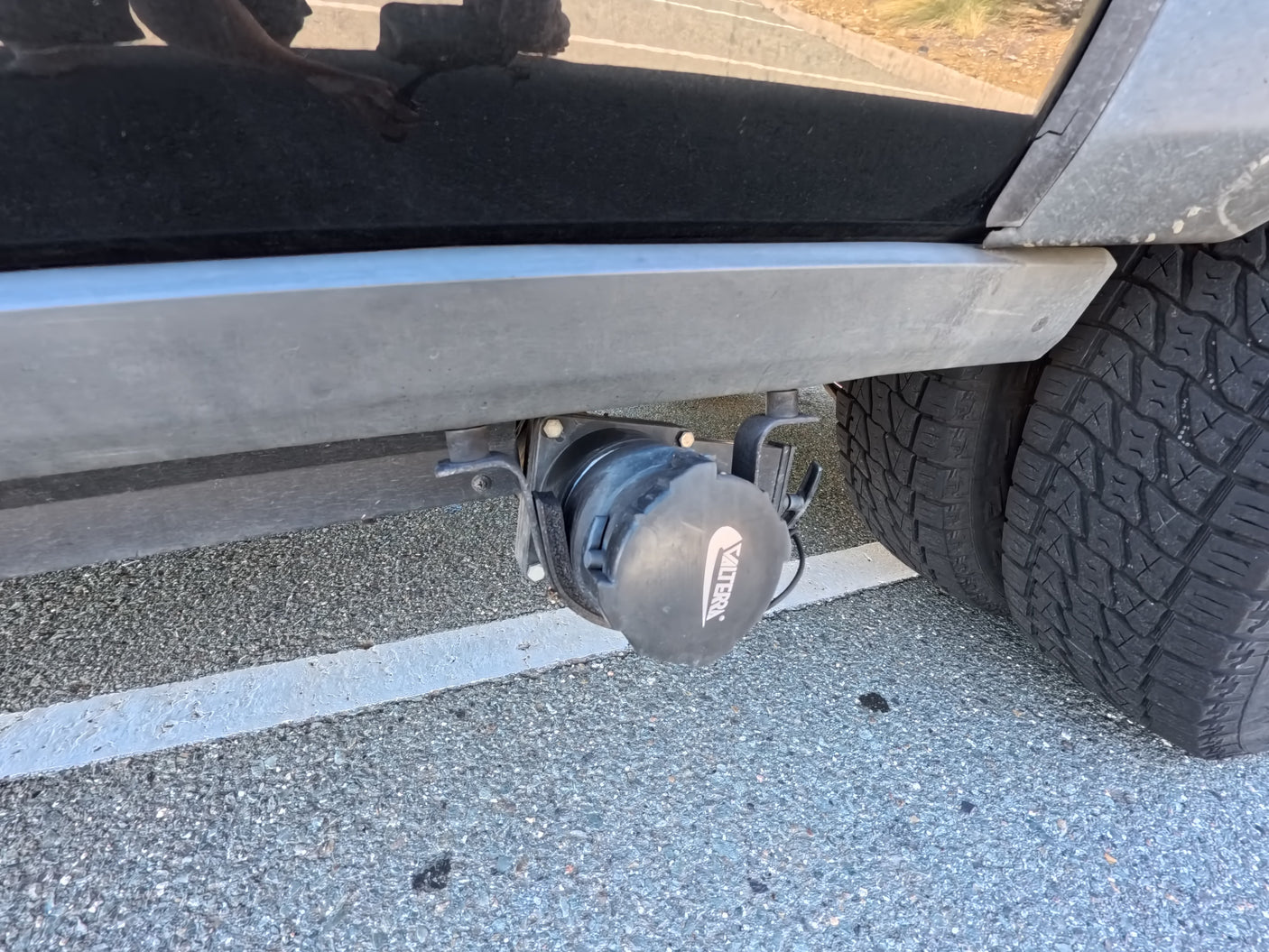 Detail of an RV rental's underside, showing a black Valterra sewer cap mounted under the chassis near dual tires on asphalt.