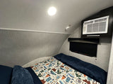 Cozy RV rental bedroom featuring a bed with a camping-themed blanket, wall-mounted AC unit, and smoke detector.