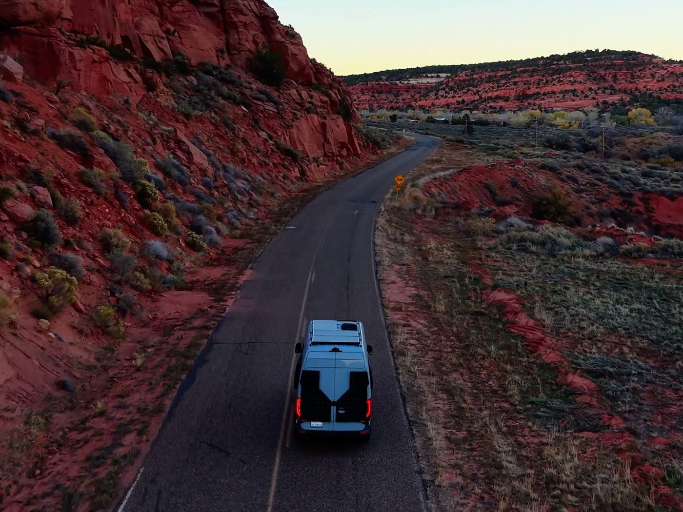 A blue RV rental van drives on a winding road through a scenic red rock desert with sparse plants under a pale sky.|Aerial view of a blue RV rental van on a desert road — photo 1