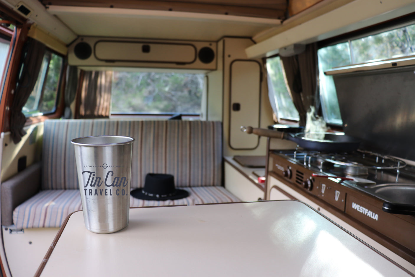 Inside a cozy Westfalia rv rental camper van: table with a metal cup, striped bench, and kitchen with a pan.