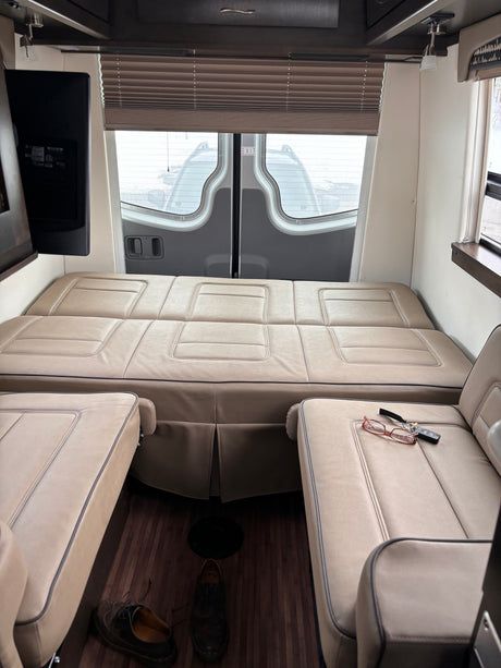 Interior of an RV rental: a light brown leather modular bed, two armchairs, a TV, and rear windows, perfect for trips.