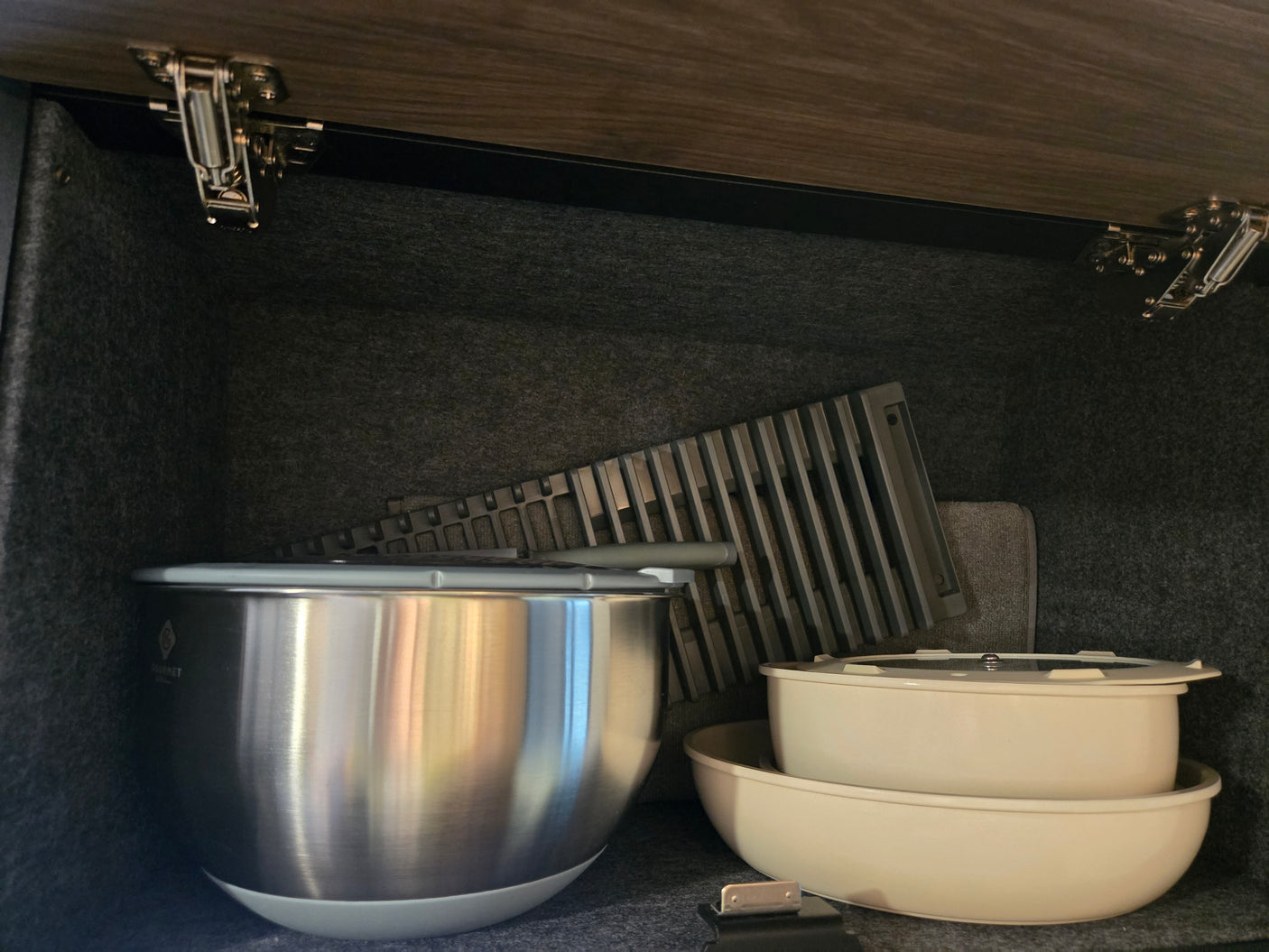Organized RV rental kitchen cabinet with a stainless steel bowl, stacked cream dishes, and a drying mat. Felt-lined for secure storage.