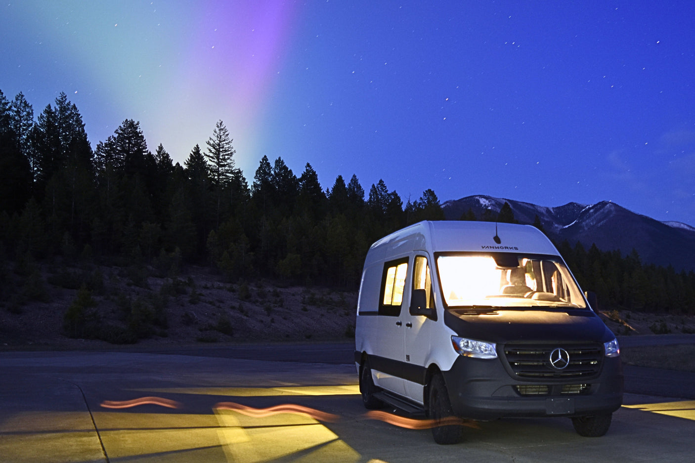 RV rental: Cozy camper van under a starry, aurora-lit night sky. Mountains and trees complete the scenic view.