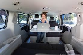 Inside a converted RV rental minivan, a woman works on a laptop at a table. It's a compact mobile office with a bed, ideal for remote work.