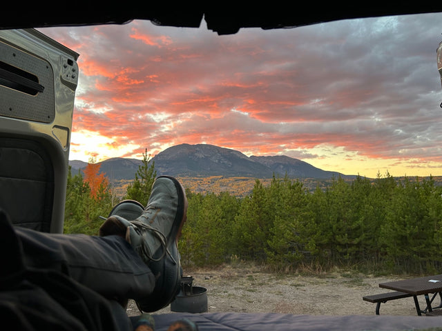  revealing a cozy interior as a person enjoys the vast canyon vista.|View from an RV rental looking out at a stunning sunset over mountains & fall trees. Legs with shoes rest inside