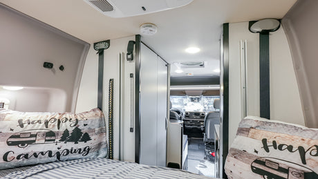 Inside a modern RV rental: From the rear bed with 'Happy Camping' pillows, looking into the spacious cockpit.