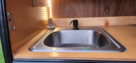 A compact wooden RV rental kitchen featuring a stainless steel sink with a soap dispenser and a brush, lit by LED strips.